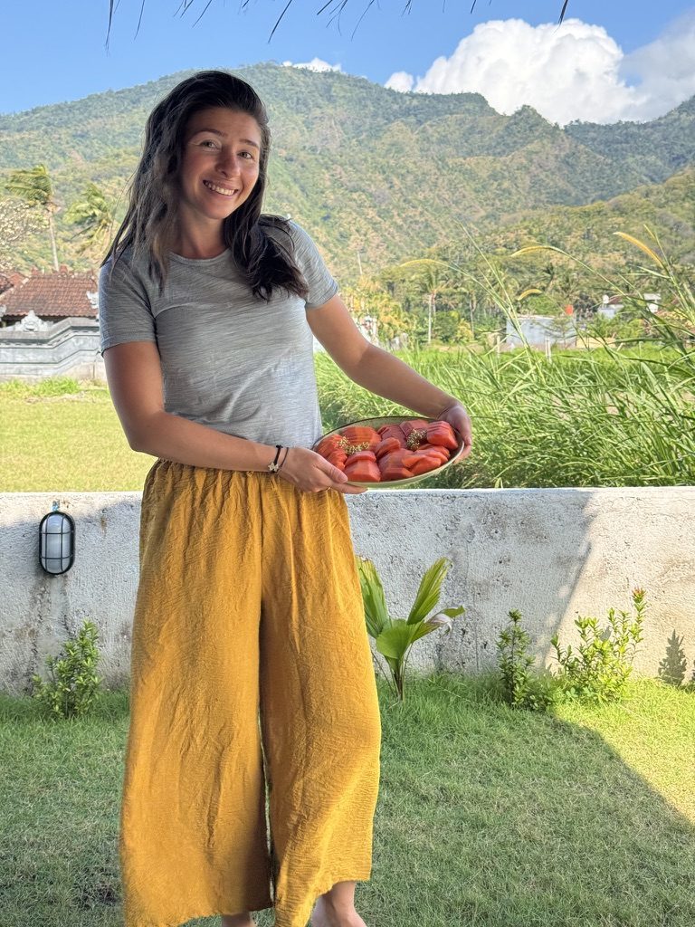 Anca smiling outdoors in Bali, holding a bowl of tropical fruit with lush green mountains behind her