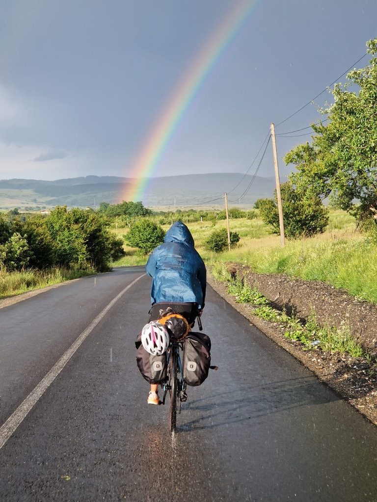 Anca cycling alone on a rain-soaked road in Alba-Iulia, Romania with a full rainbow arching overhead