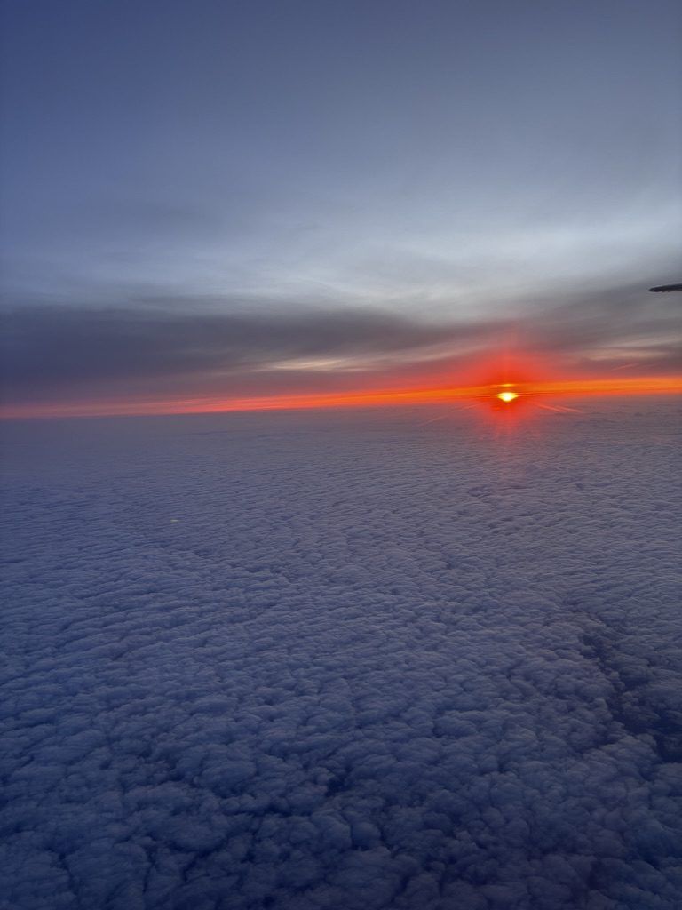 Sunrise viewed from an airplane window over the Atlantic, pink and orange light across the clouds