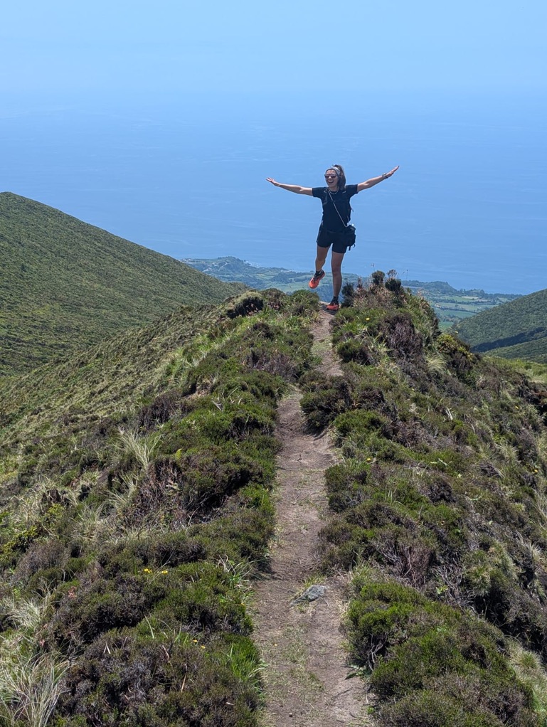 Anca on a ridge in the Azores with arms wide open, island spread out below