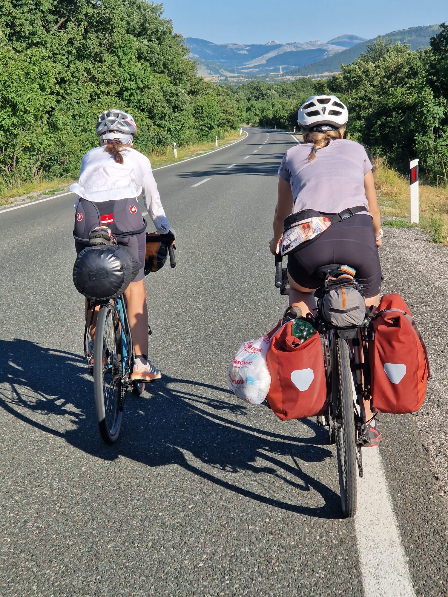Two cyclists with loaded panniers riding side by side on a rural road, hills in the background