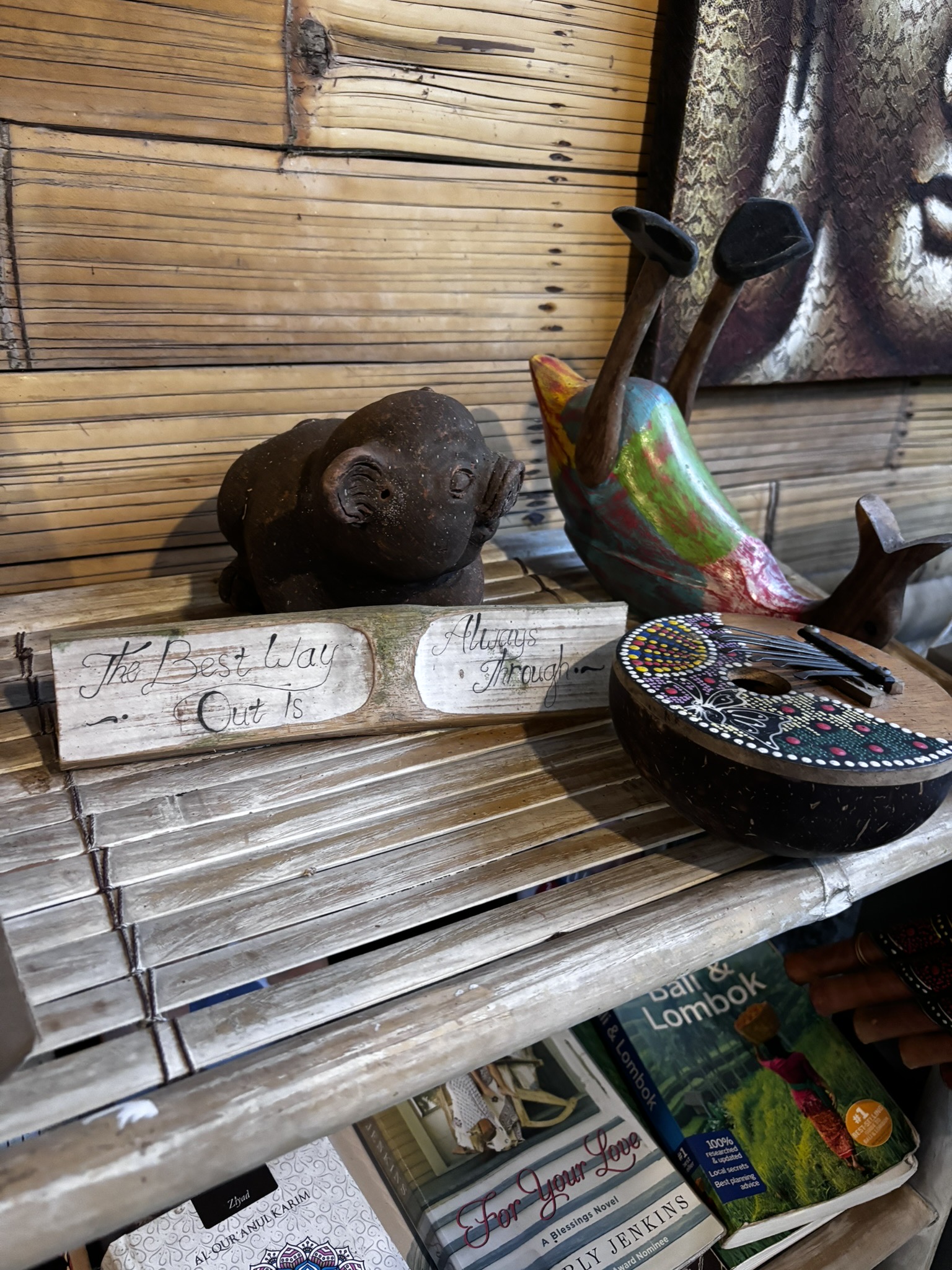 A carved wooden sign reading 'The Best Way Out Is Always Through' on a Balinese gift shop shelf, surrounded by stone figurines, a painted duck, and a decorated mask