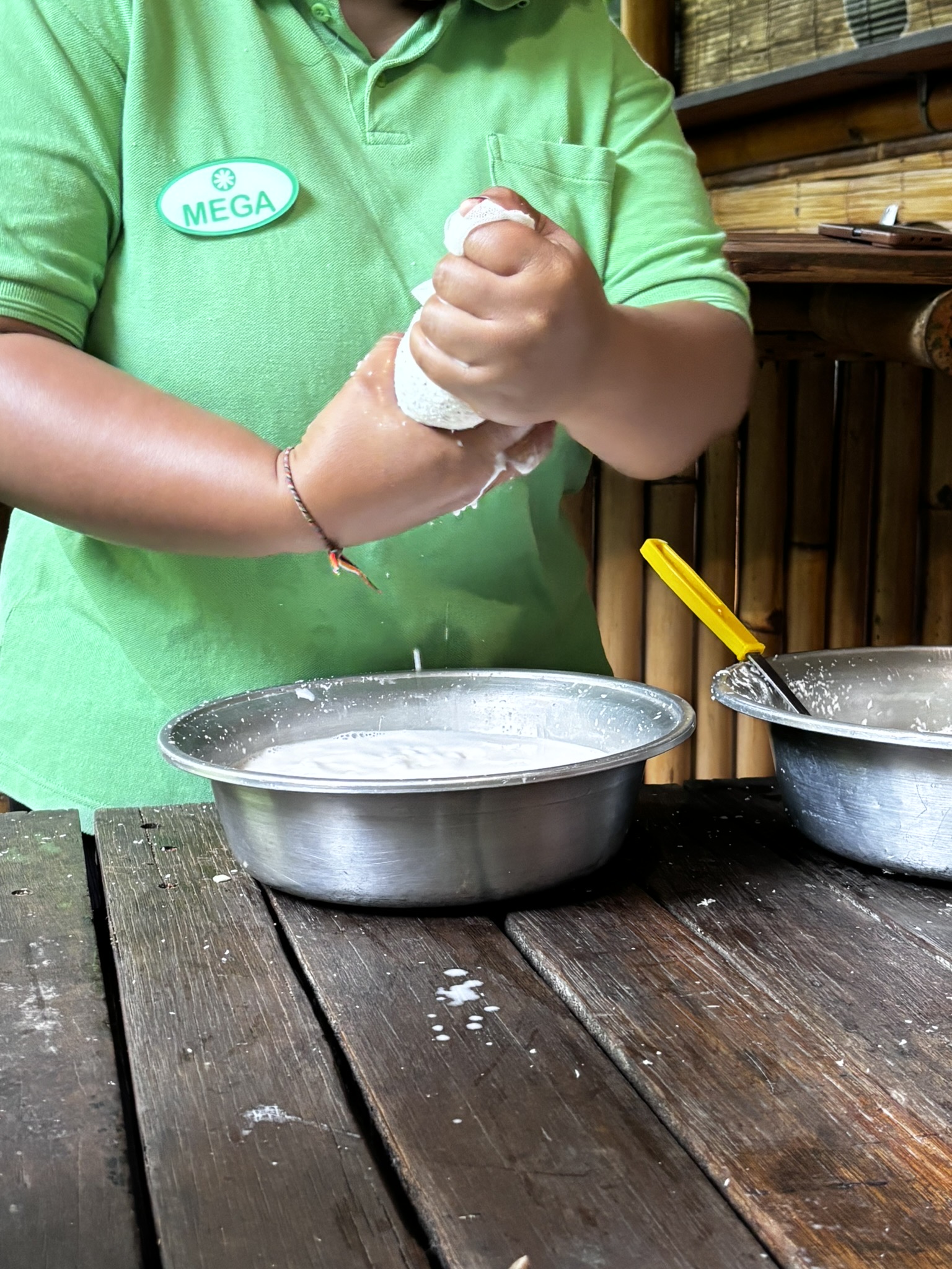 Close-up of Mega, a Balinese workshop instructor in a green polo shirt, squeezing grated coconut flesh in both hands over a metal bowl to extract coconut milk