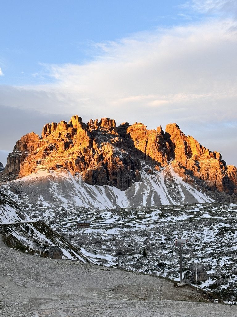 Massive Dolomite massif glowing deep orange in alpenglow light, fresh snow on lower slopes, snowy path in foreground