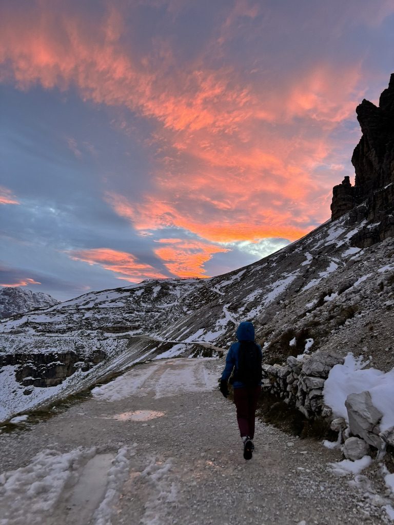 Anca walking on a snowy mountain path under a violent orange and red sunset sky in the Dolomites