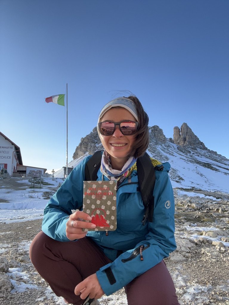 Anca holding up a Dolomiti hiking passport at a rifugio, Italian flag behind her, rocky peaks and snow beyond