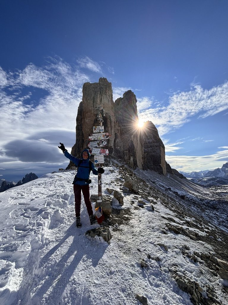 Anca at a trail signpost in deep snow, arms raised, with the iconic peaks of Tre Cime di Lavaredo rising dramatically behind her in the sun