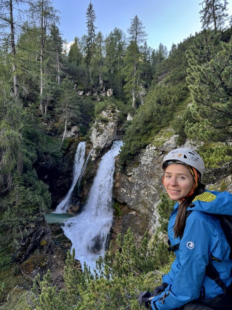 Anca smiling in a climbing helmet next to a tall waterfall, pine forest behind, Dolomite rock face