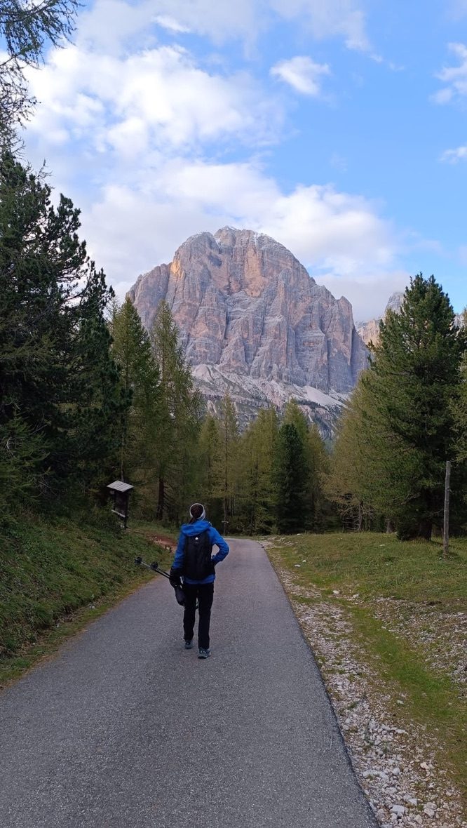 A hiker walking away down a path framed by larch trees, a massive vertical Dolomite tower filling the sky ahead