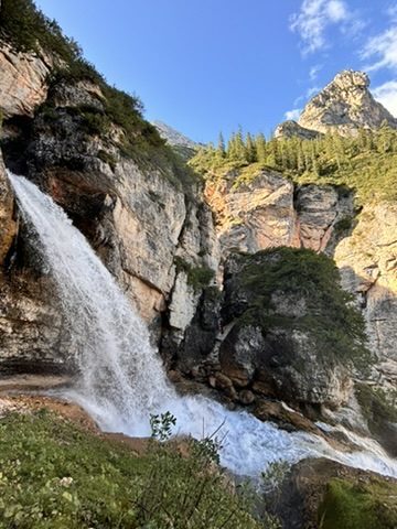 A powerful waterfall cascading down orange and pink Dolomite rock face, green pine forest above, blue sky