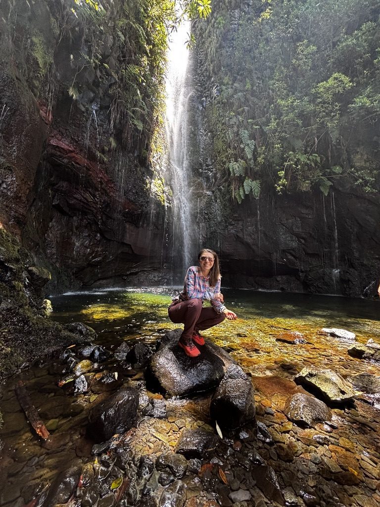 Anca sitting on a rock in front of the 25 Fontes waterfall, surrounded by lush green vegetation, light filtering through the jungle canopy