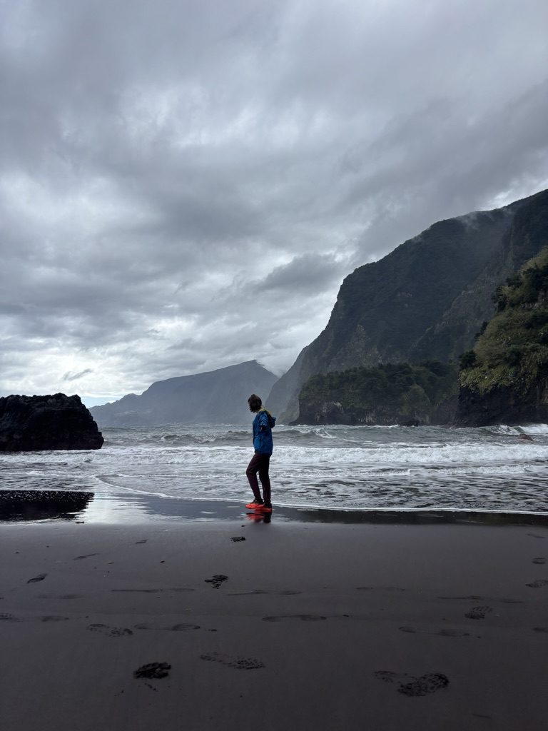 Anca standing alone on a dramatic black volcanic sand beach, stormy waves, sheer green cliffs behind in fog