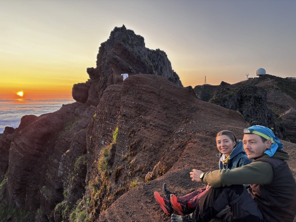 Anca and a friend sitting on volcanic rock at Pico Arieiro summit, sun setting on the horizon below the cloud layer, warm orange light