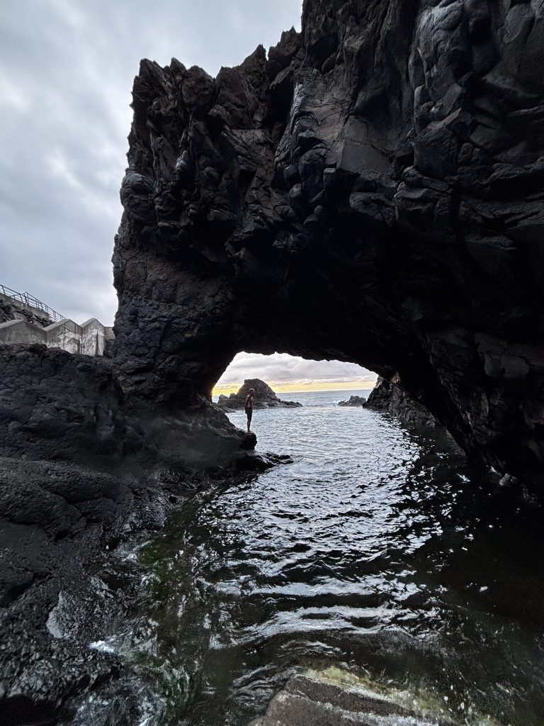 Silhouette of a person standing through a massive natural volcanic rock arch, ocean and sunset beyond, dark wet rock in foreground