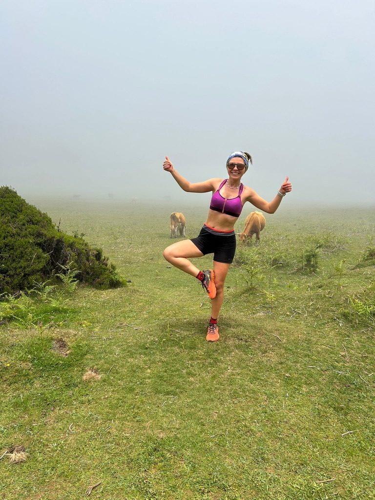 Anca standing on one leg in a yoga pose, thumbs up, in fog on a green plateau, cows grazing behind her