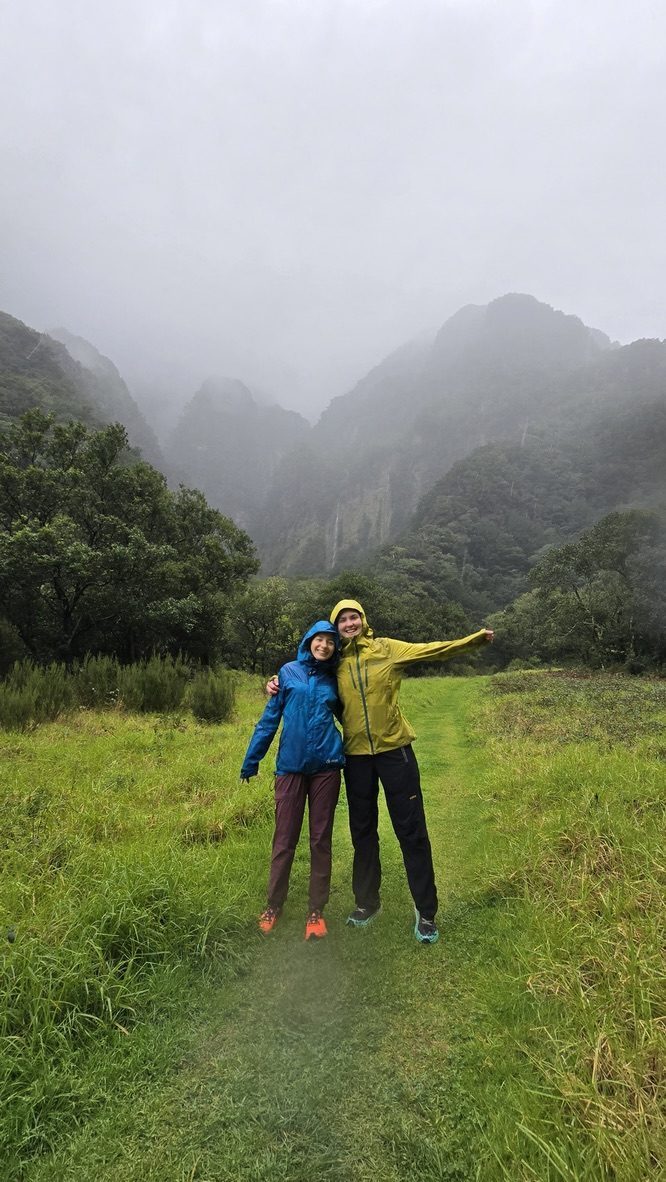 Anca and a tall friend in rain gear, both smiling broadly, standing in a green valley with dramatic misty cliffs and a waterfall behind