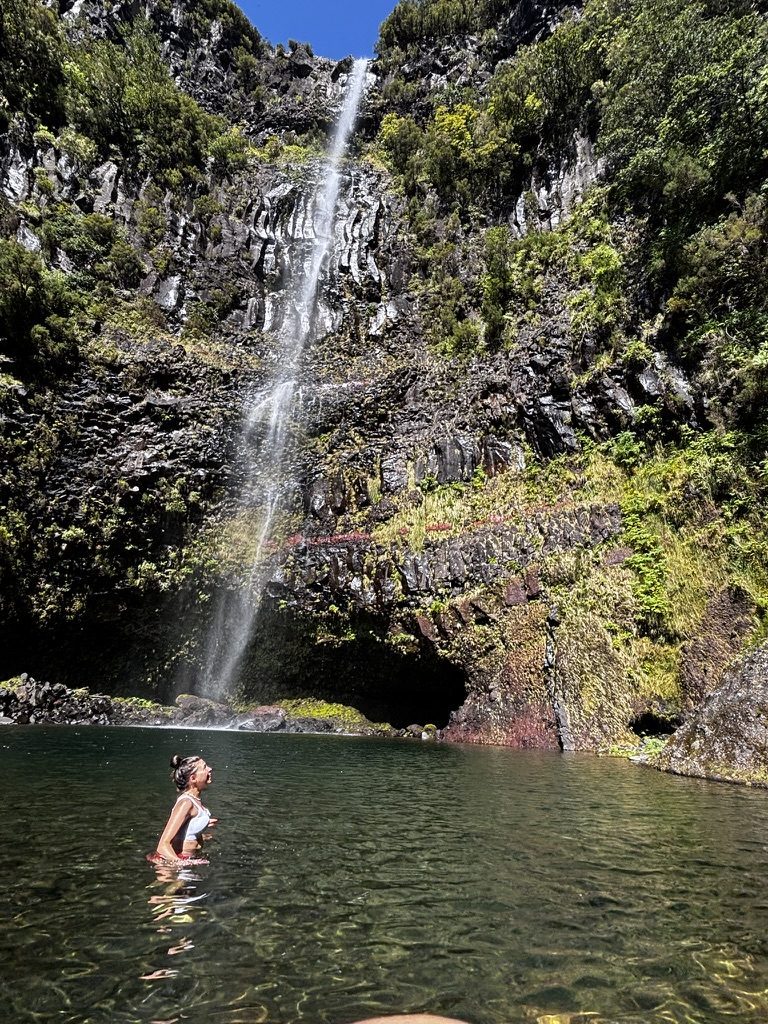 Anca standing waist-deep in a natural pool at the base of a tall waterfall, sunshine overhead, lush green tropical vegetation