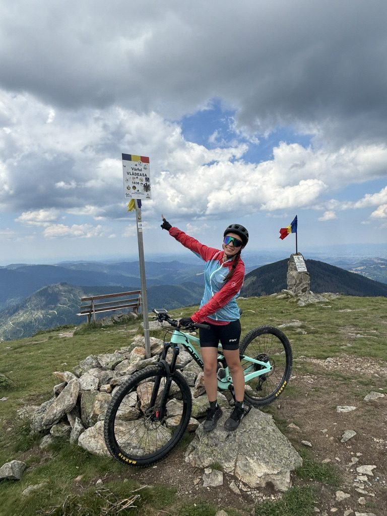 Anca at Vârful Vlădeasa summit sign at 1835m, red and blue jacket, Orbea beside her, Romanian flag and dramatic clouds behind