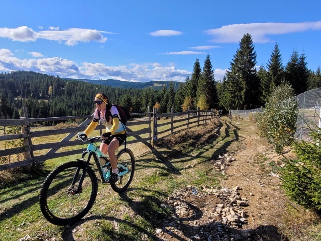 Anca riding the Orbea alongside a wooden fence on a trail, pine forests and autumn colours behind
