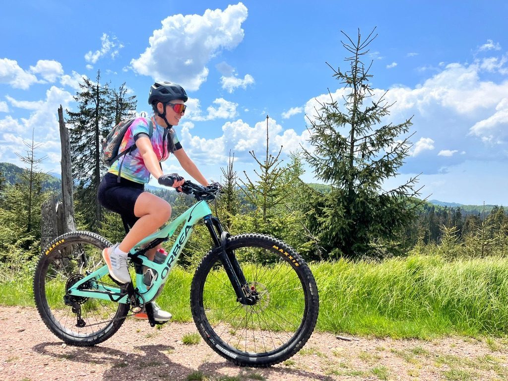 Anca on the mint Orbea on a gravel trail, conifer forest and mountains behind, blue sky with clouds