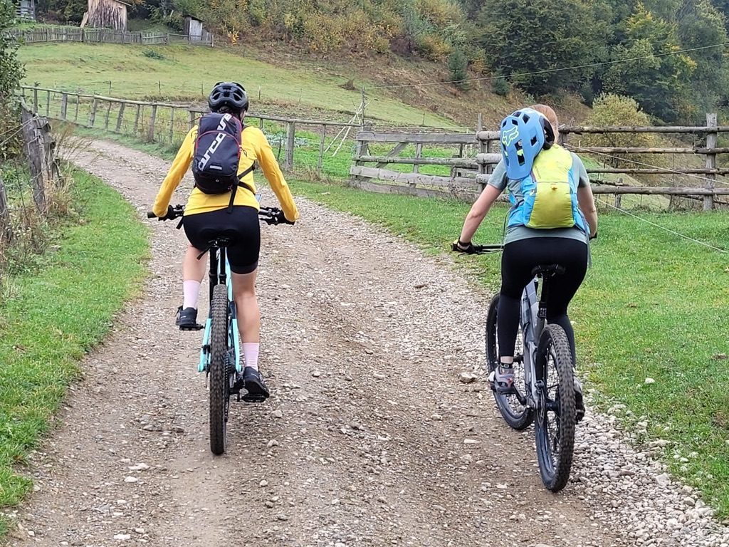 Two women riding MTBs on a gravel path through a valley, wooden fences, autumn foliage