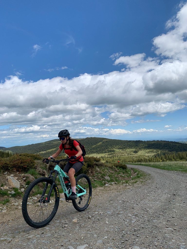 Anca riding the Orbea on a gravel ridge road, vast open mountain landscape behind, big sky