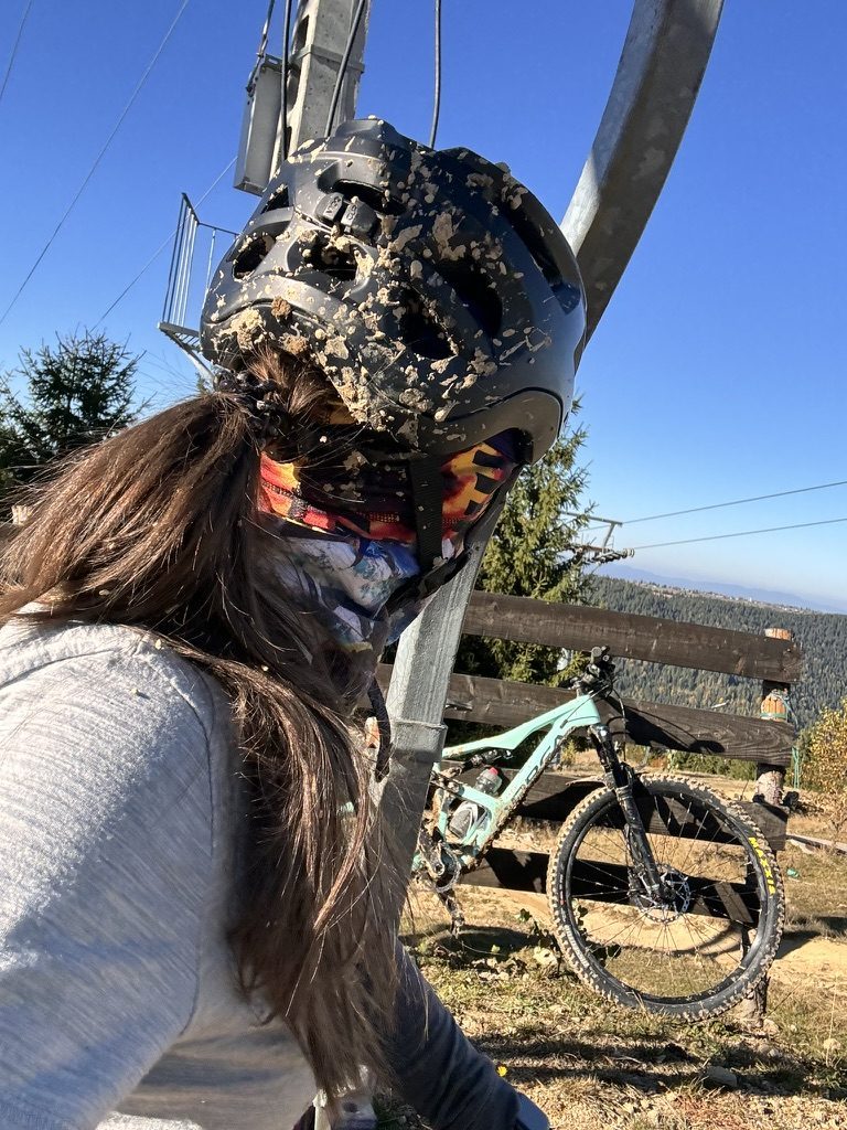 Anca's helmet and face completely caked in mud after a fall, blue sky behind, Orbea bike visible in background