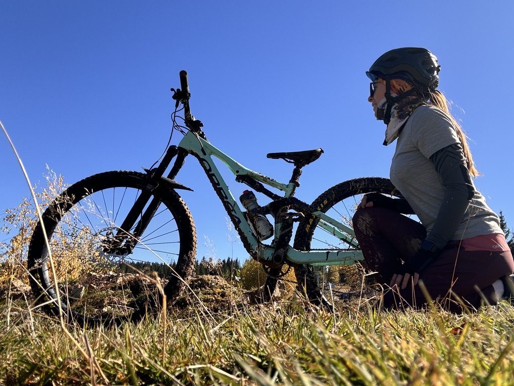 Anca kneeling beside her Orbea MTB in autumn grass, blue sky