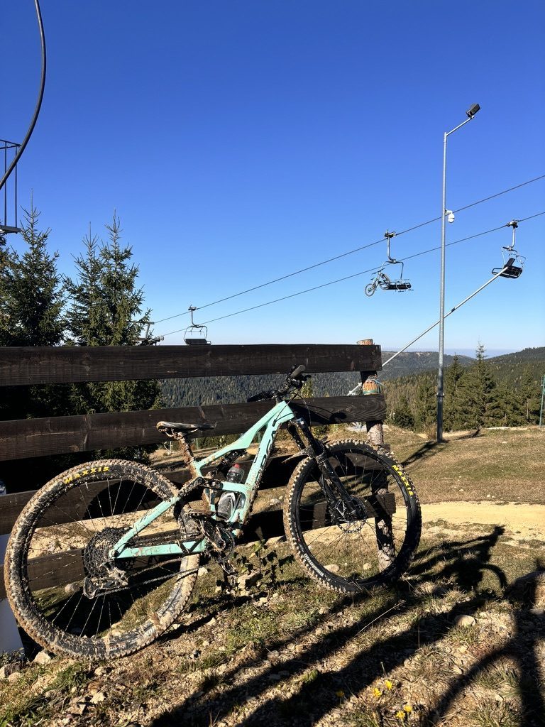 Mint Orbea MTB leaned against wooden fence at top of chairlift, caked in mud, blue sky and forest behind