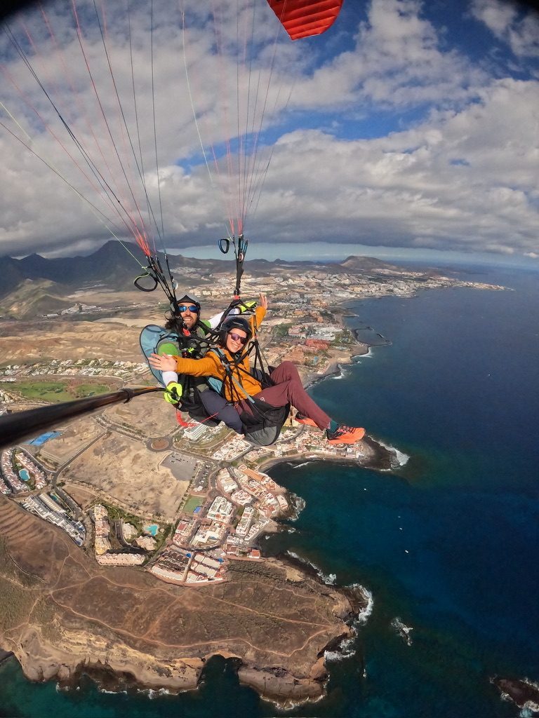 After the paragliding landing — Anca and pilot in the landing field, Tenerife coast behind them, the joy is real