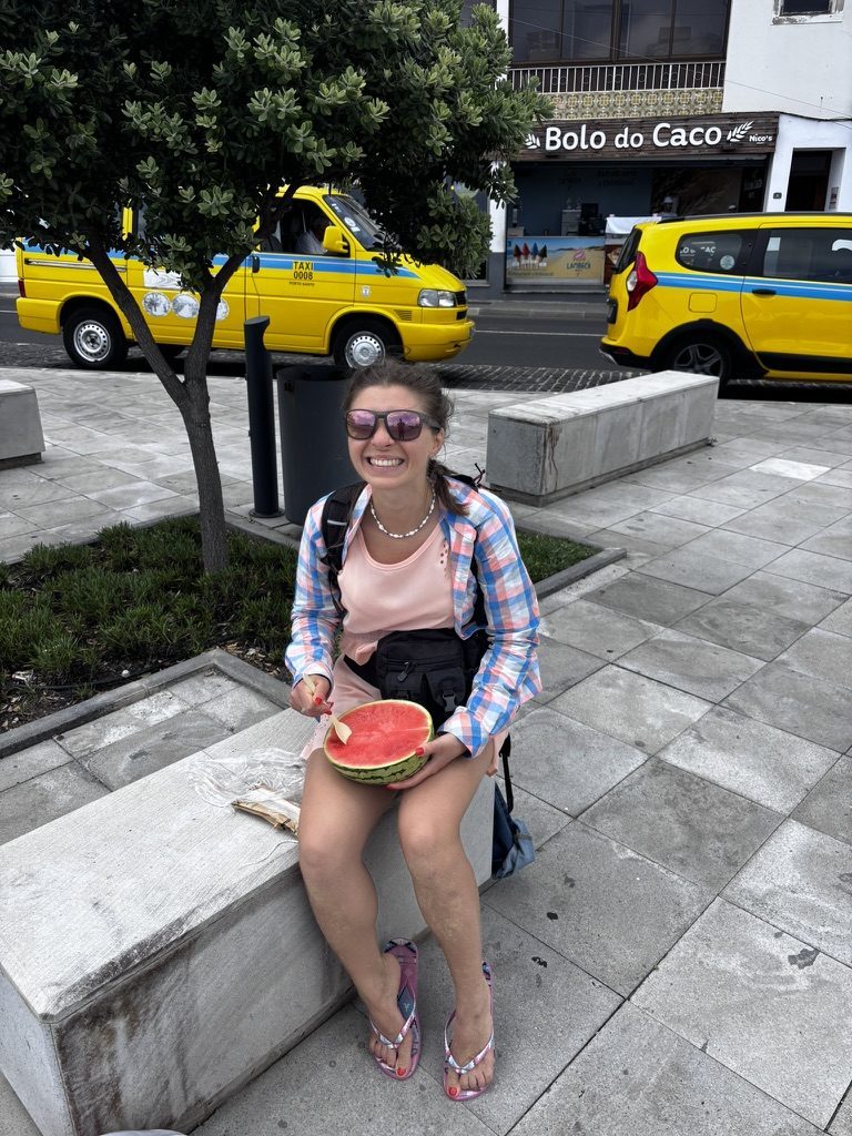 Anca sitting on a low wall outside eating half a watermelon with a spoon, Porto Santo yellow taxis behind her, laughing