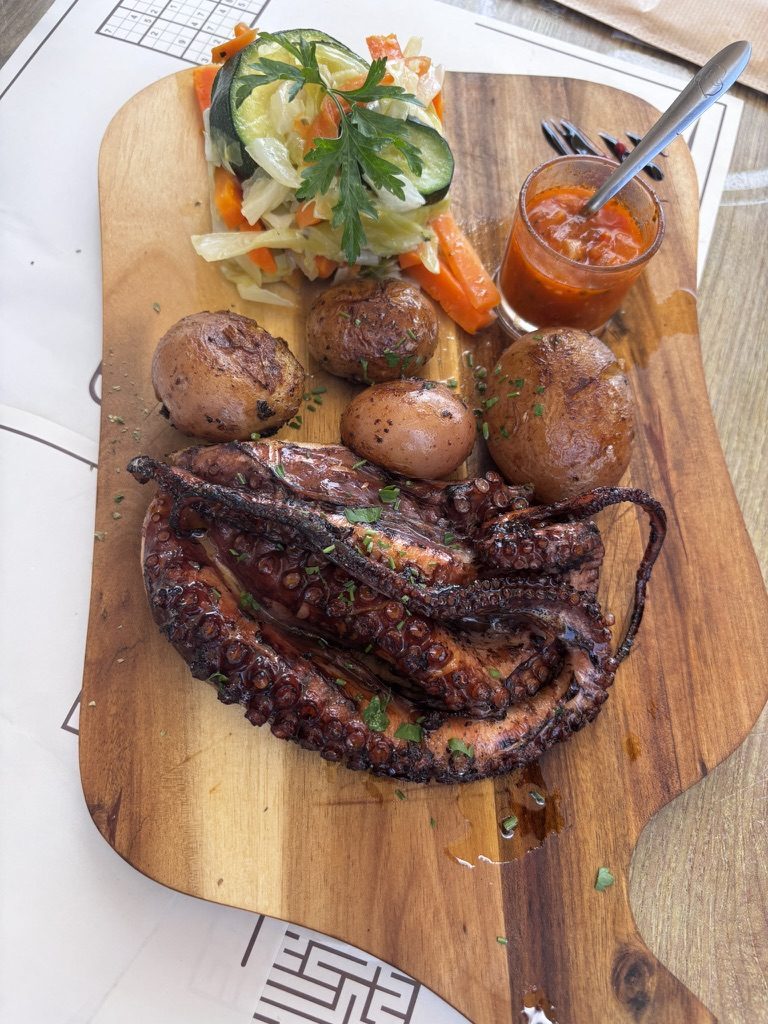 A whole grilled octopus on a wooden serving board with roasted potatoes, salad and tomato sauce