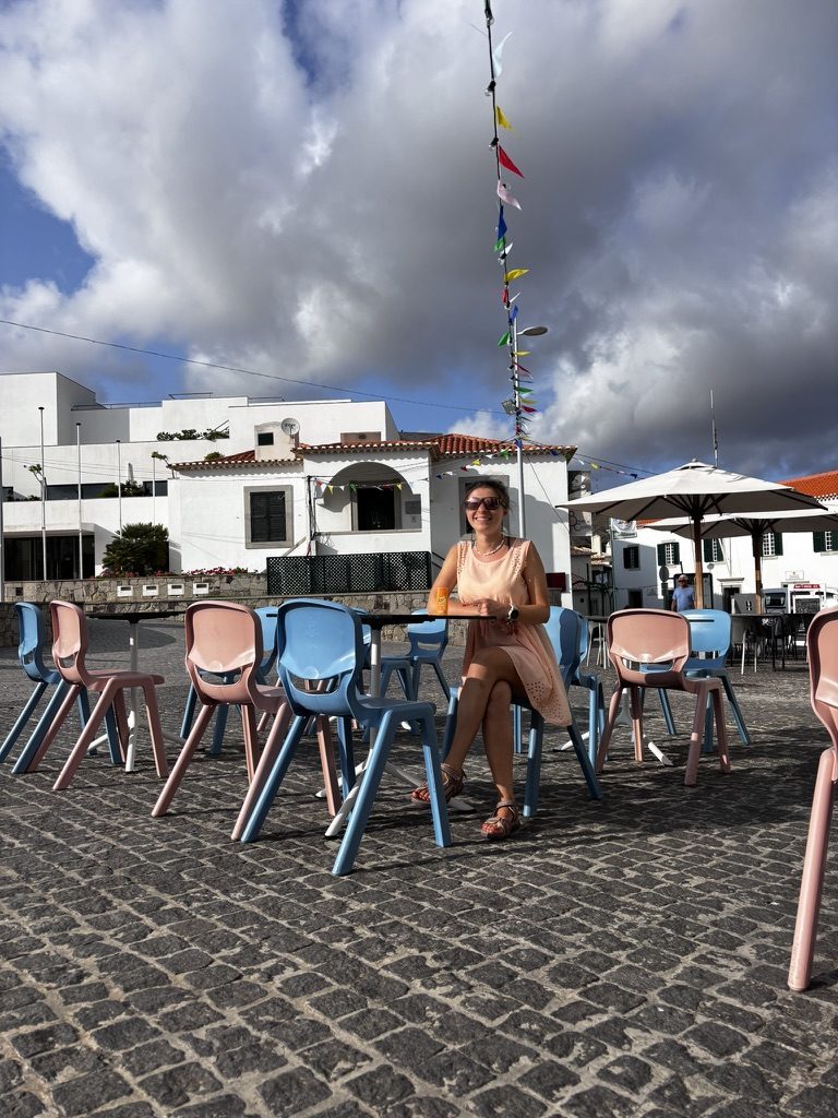 Anca sitting alone at a table in a colourful outdoor cafe in Porto Santo's village square, pastel chairs, festival bunting above