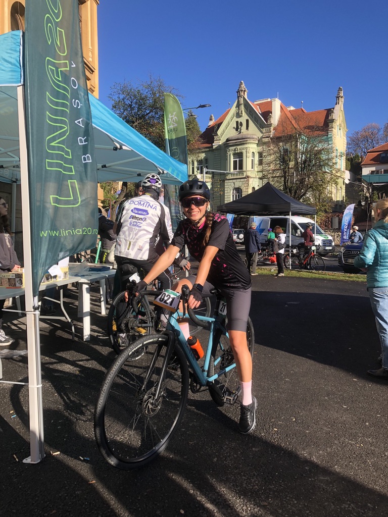 Anca at the Linia 20 Brașov race start, smiling at the camera with race number 021 on her blue bike, Art Nouveau buildings and event tents behind her in the autumn sun
