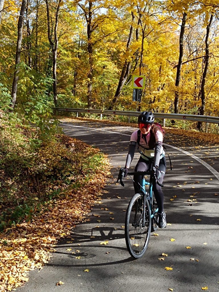 Anca cycling alone on a winding forest road deep in autumn colours — golden and orange leaves cover the trees and the tarmac, a curve ahead disappearing into the forest