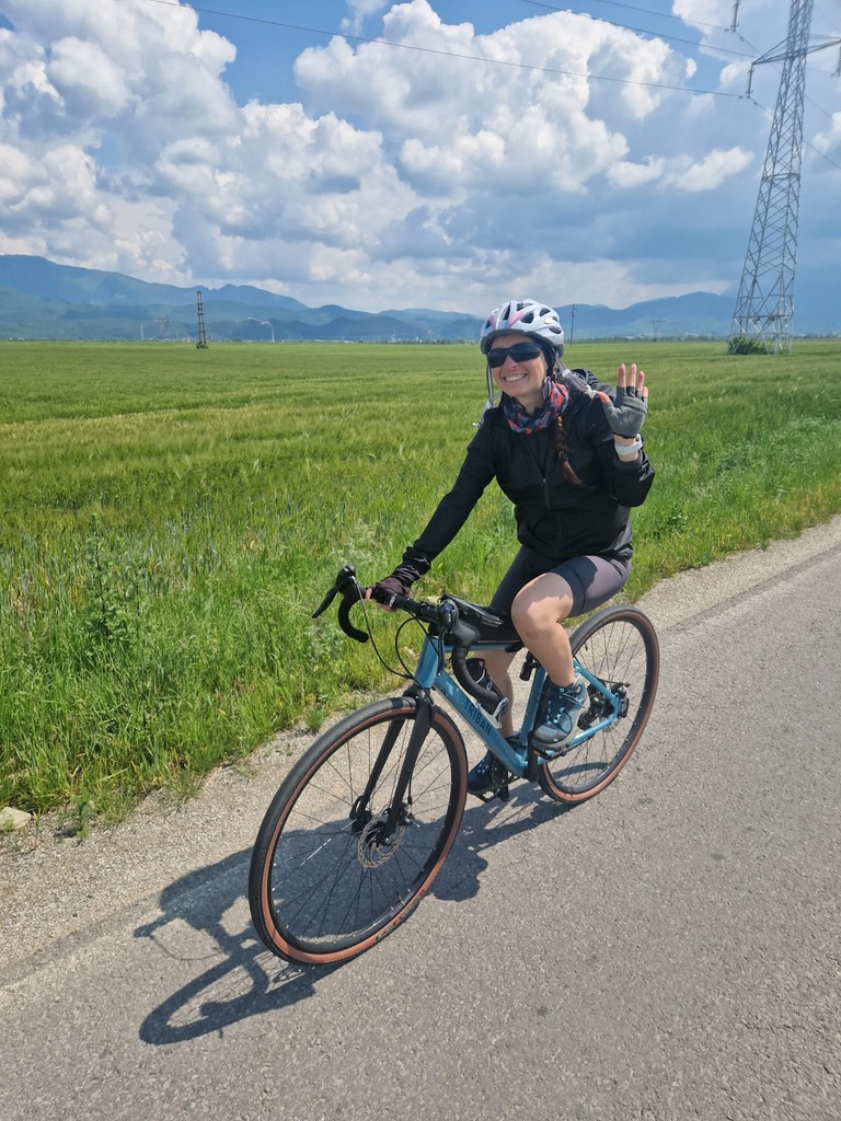 Anca on her blue gravel-road bike on a country lane near Brașov, waving at the camera, green fields stretching behind her, the Carpathian mountains on the horizon under a dramatic cloudy sky
