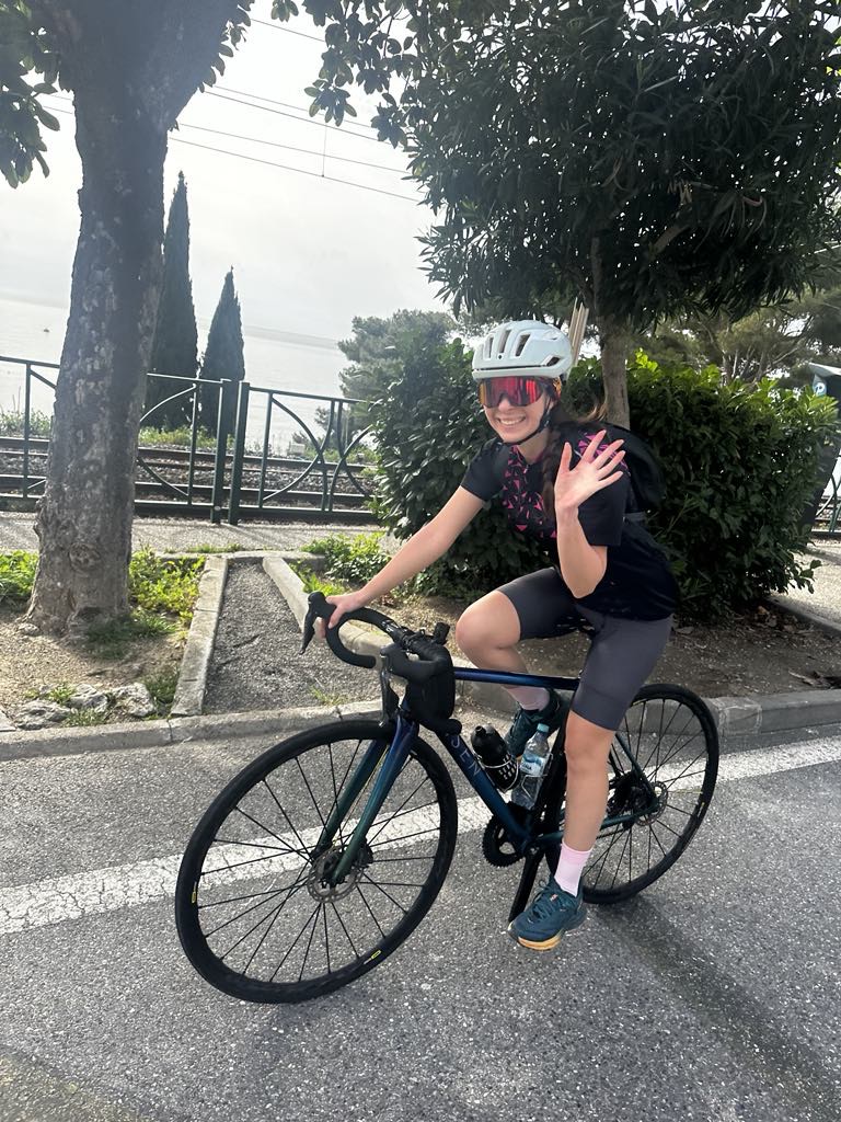 Anca on a dark road bike in Nice, waving at the camera, cypress trees and Mediterranean architecture behind her, white helmet and pink sunglasses