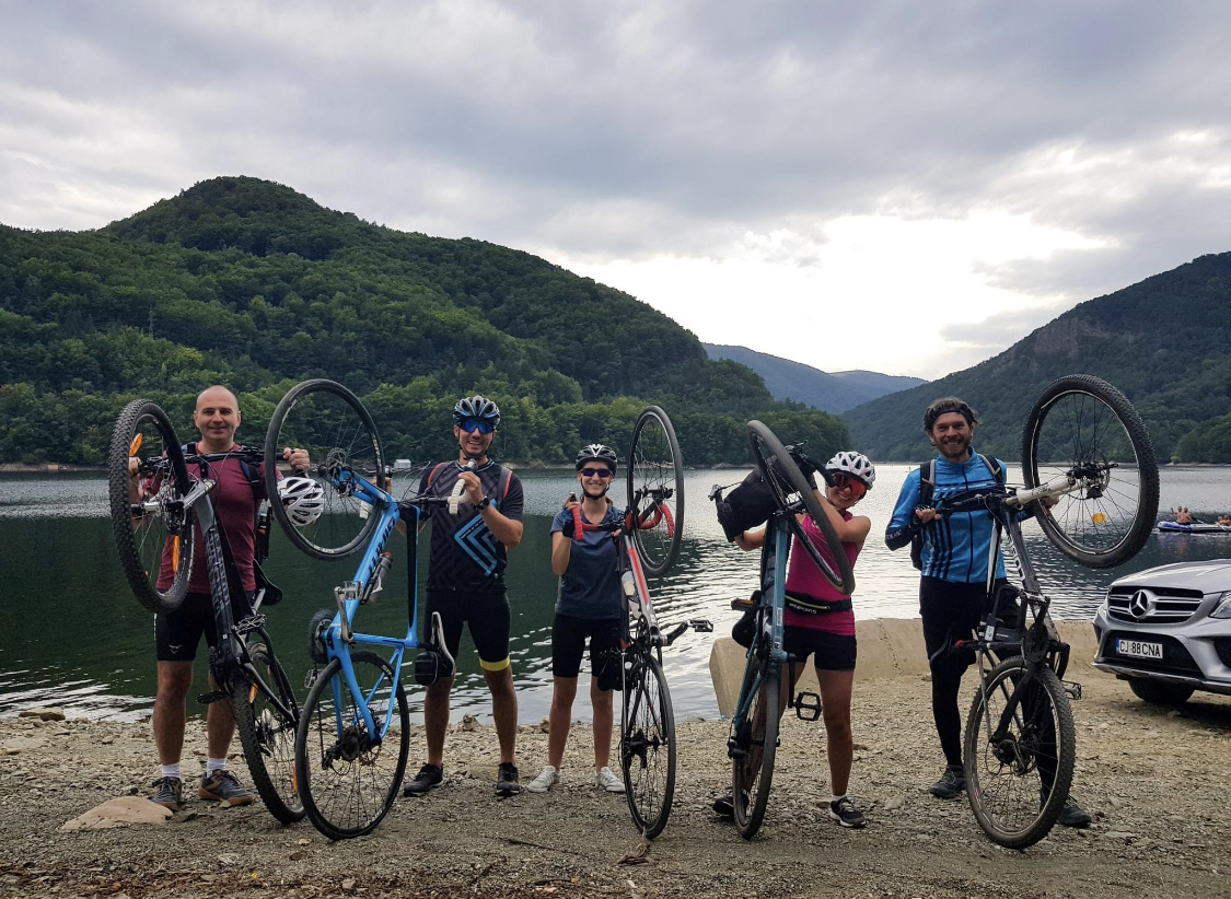 Five cyclists holding their bikes triumphantly overhead at the edge of a mountain lake in Romania's Apuseni region — green forested hills reflected in the grey water behind them