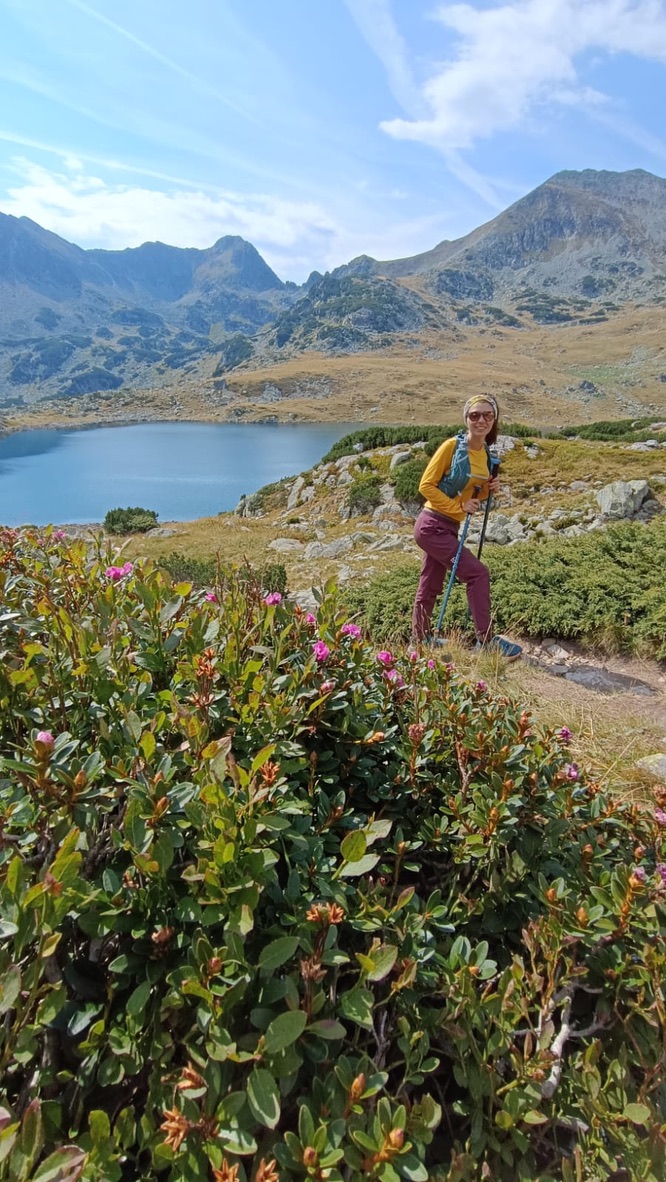 Anca smiling beside a glacial lake in the Retezat, rhododendron flowering in the foreground, dramatic rocky peaks and blue sky behind her