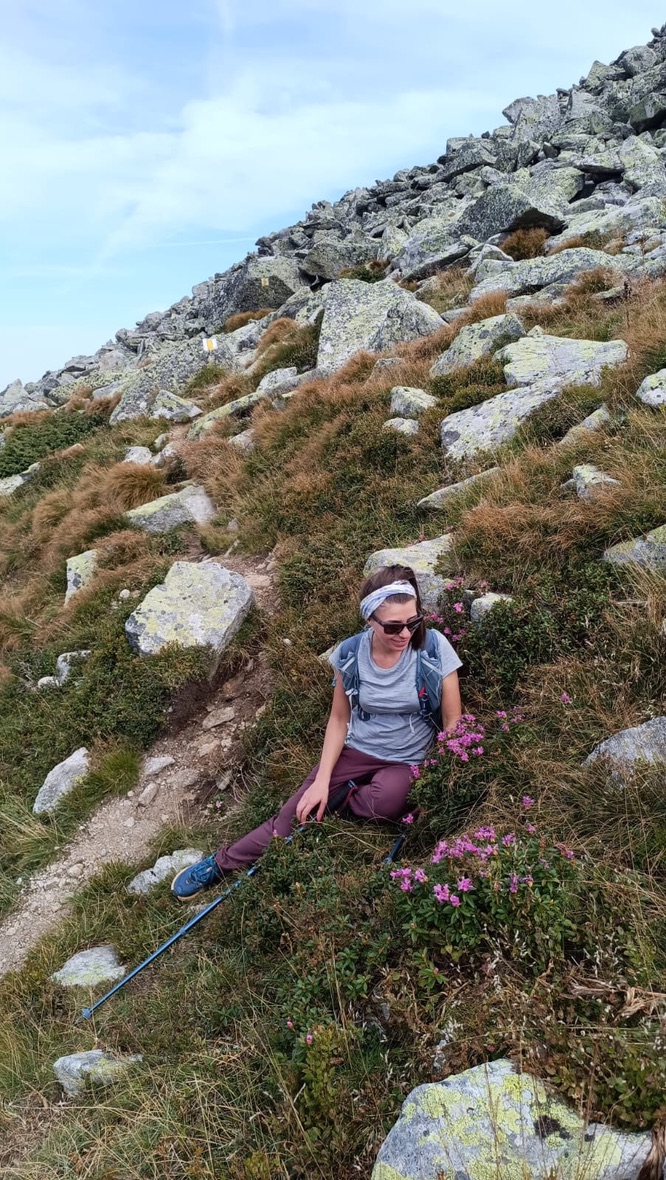 Anca sitting on a steep rocky slope in the Retezat, surrounded by small pink rhododendron flowers blooming between the rocks, resting mid-hike
