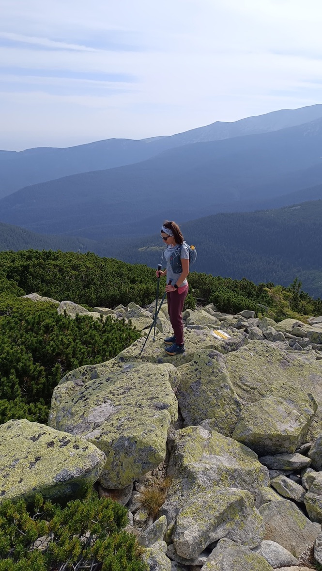 Anca standing on large lichen-covered rocks on the Retezat ridge, trekking poles in hand, the vast Romanian Carpathian ridges stretching blue into the distance behind her