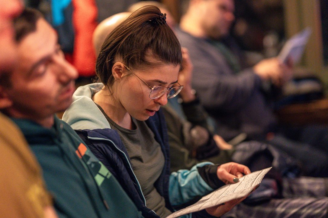 Anca reading intently from a printed programme at an indoor briefing session, surrounded by other camp participants