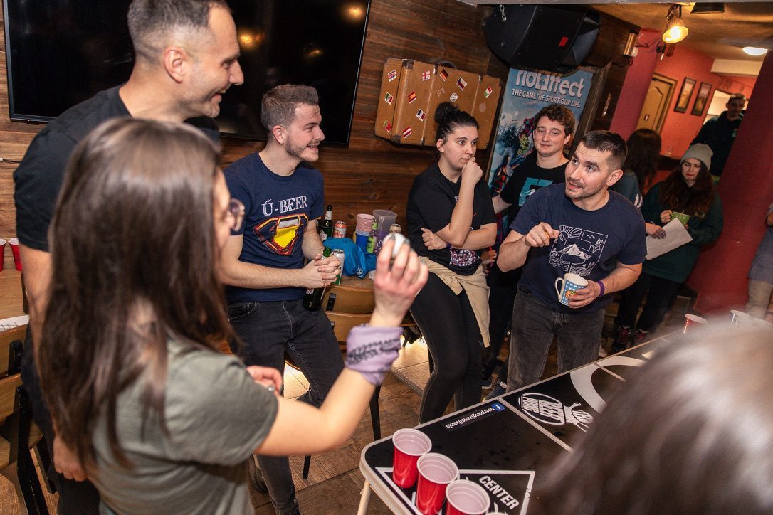 Indoor après ski scene, Anca at a beer pong table with other camp participants, laughing, ski branding on the wall
