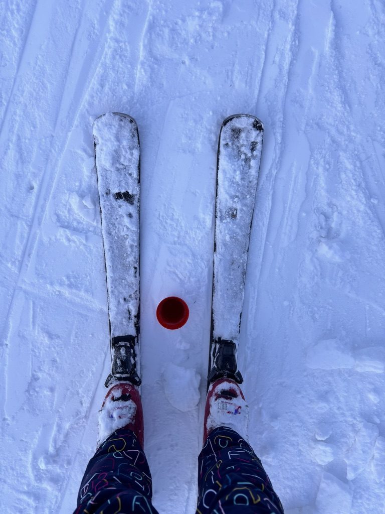 Looking straight down at Anca's ski tips on snow, a red piste marker between them, colourful ski trousers