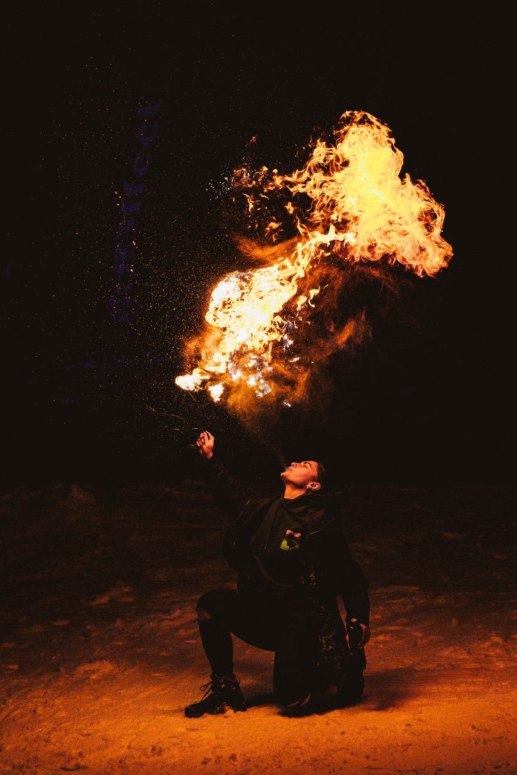 A fire breather on one knee at night, exhaling a massive fireball into the darkness, snow and sparks around them