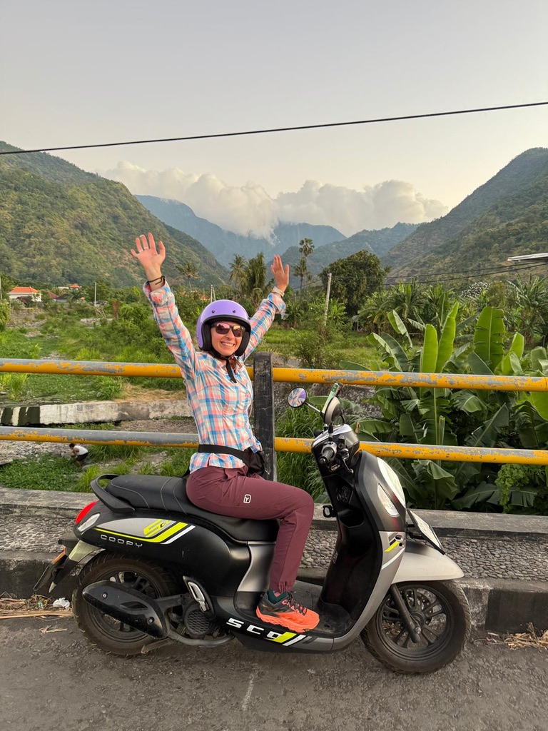 Anca on a scooter in Indonesia, arms raised triumphantly, green mountains behind her