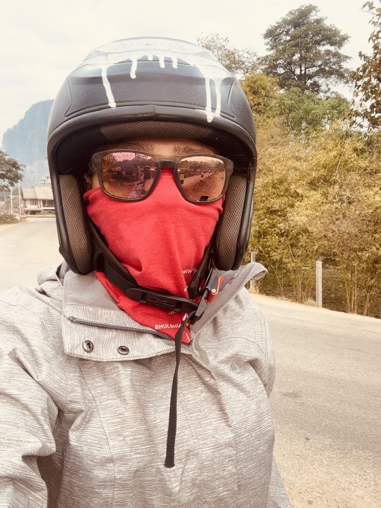 Anca in full scooter gear — black helmet, oversized sunglasses, red face mask covering everything, grey hoodie — completely unrecognisable on a road in Laos