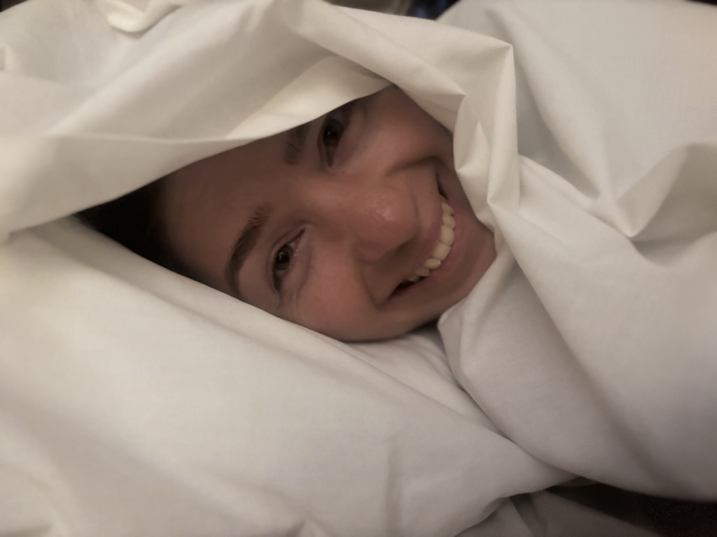 Anca's face peeking out from a cocoon of white hotel duvet, grinning enormously