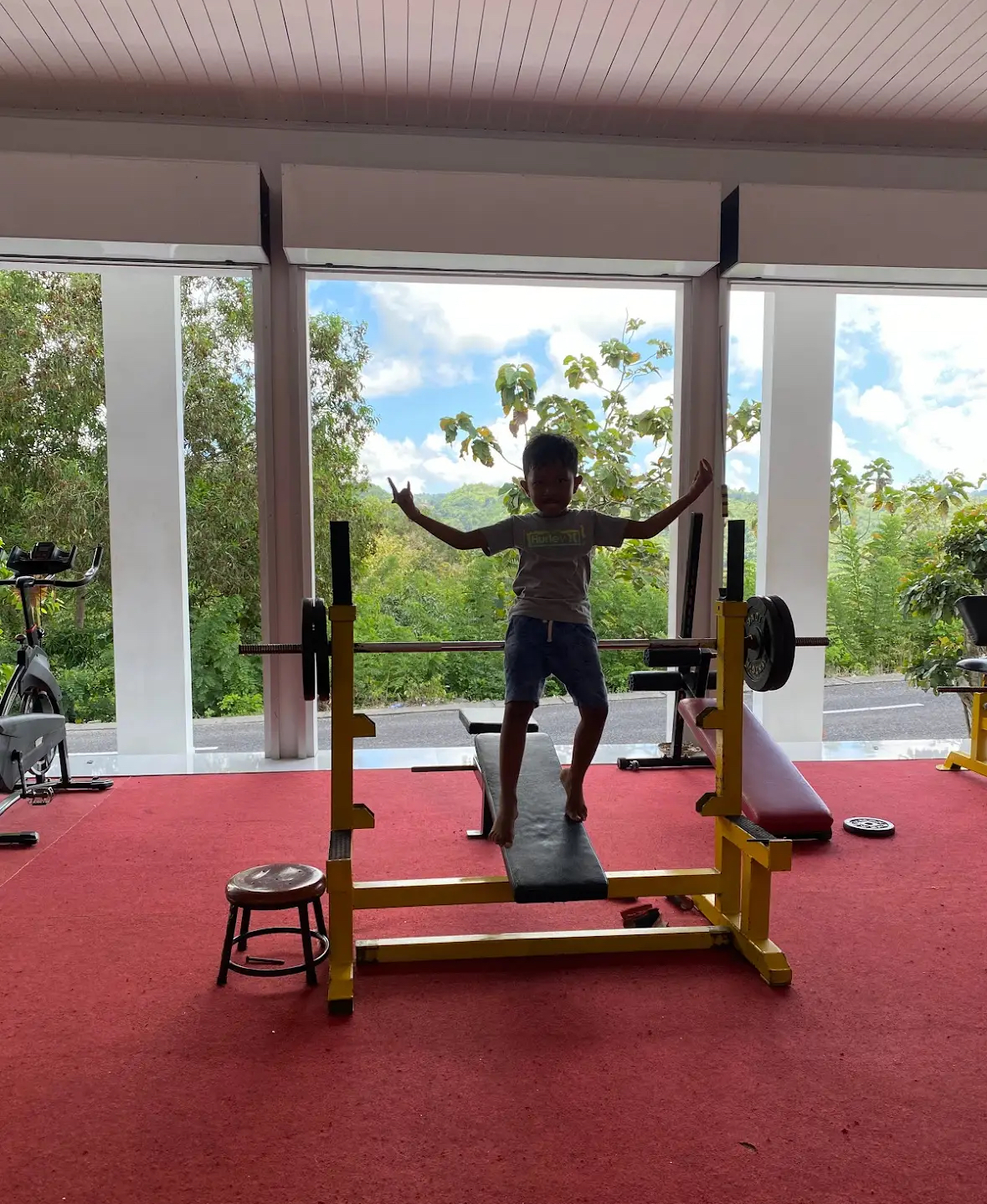 A small confident child standing triumphantly on gym equipment in a tropical hotel gym, arms wide, looking thrilled with himself