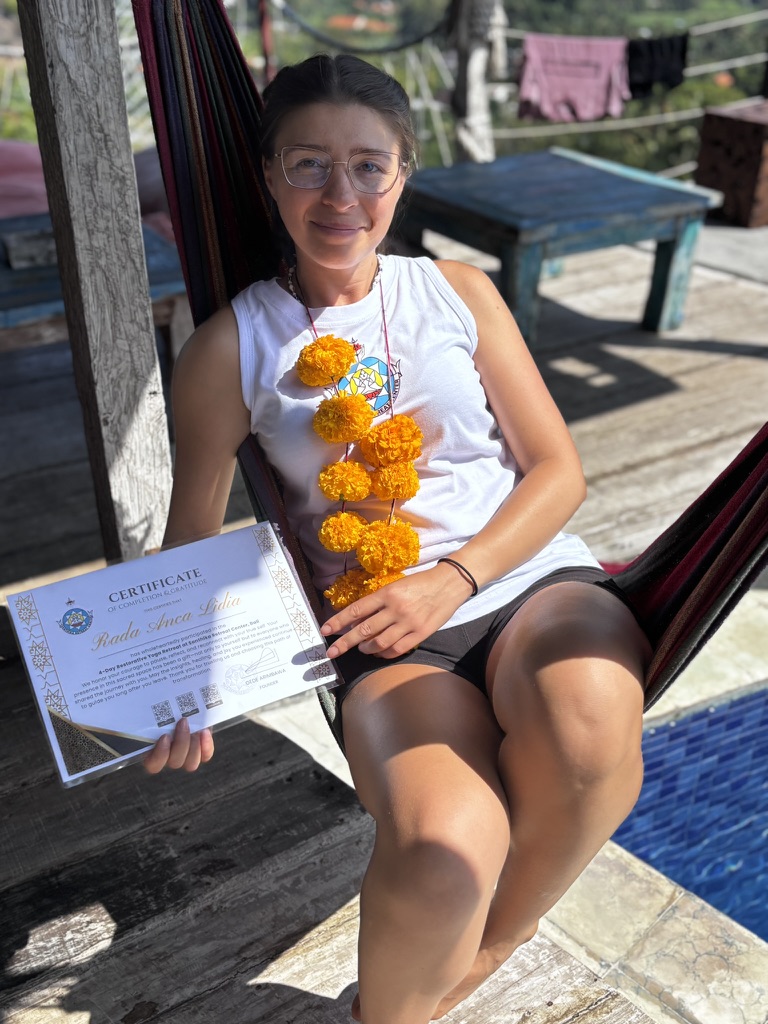 Anca sitting in a hammock in Bali, holding a yoga retreat certificate, wearing a garland of bright orange marigold flowers, looking pleased